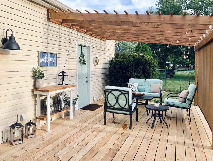 Wooden deck with pergola, outdoor seating with cushions, small side table, plants, string lights, and "welcome" sign on house wall