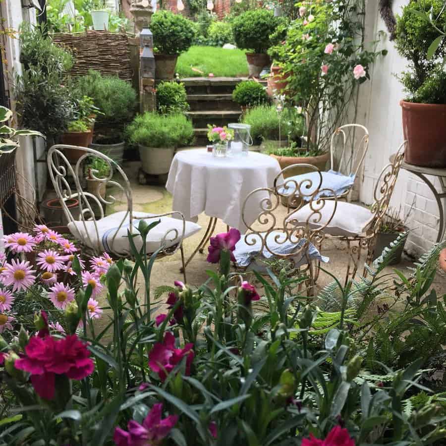 Charming garden patio with ornate metal chairs, a round table with a white cloth, surrounded by lush flowers and greenery