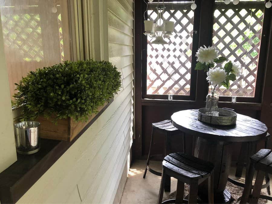 Cozy porch with a small round table, stools, potted white flowers, and a box of greenery, surrounded by wooden lattice panels