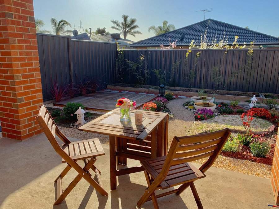 Cozy backyard patio with wooden furniture, vibrant flower beds, a small deck, and a private fence under sunny skies