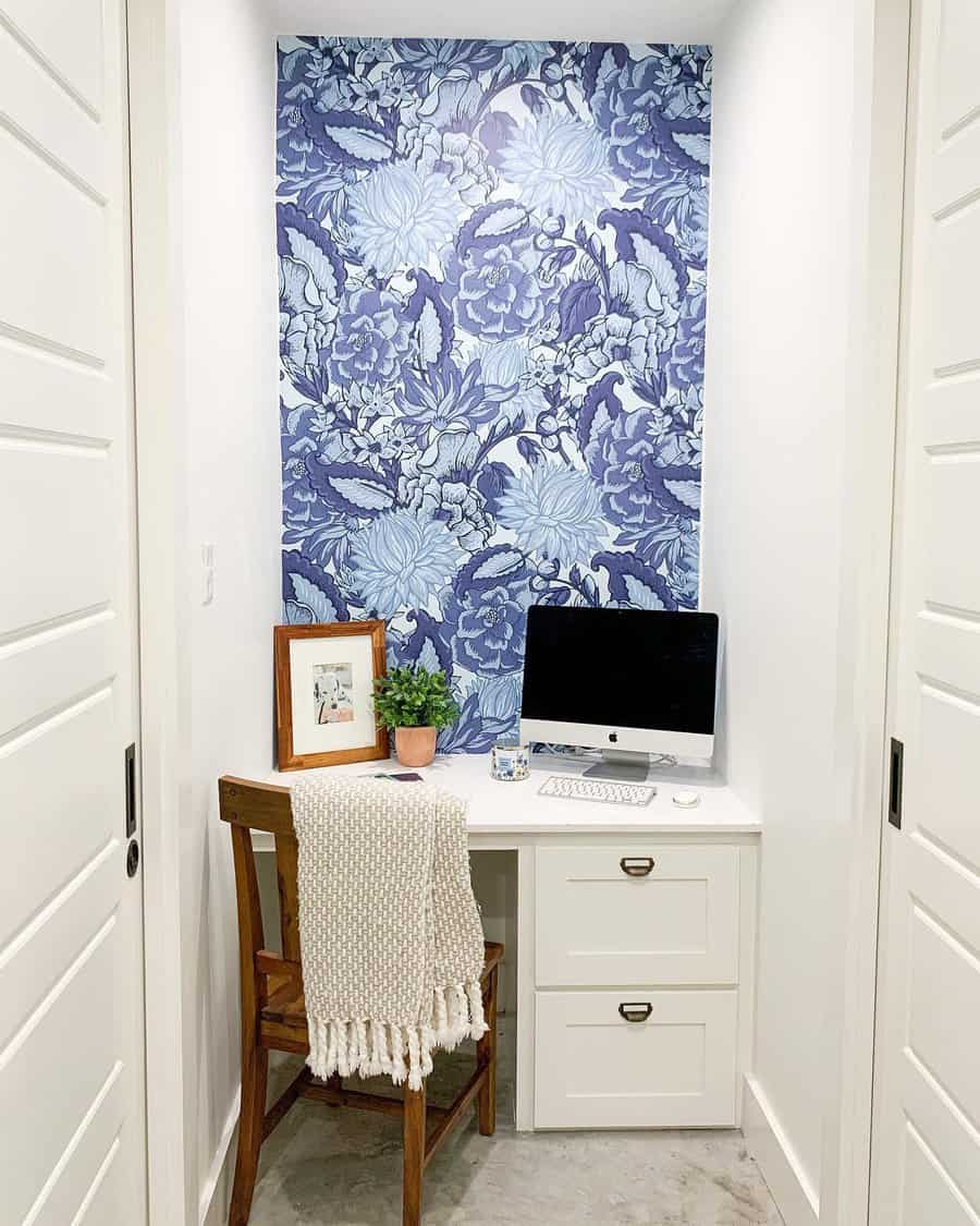 Small white desk with computer, chair, plant, and framed pictures. Blue floral wallpaper accent behind. Cozy office nook setup