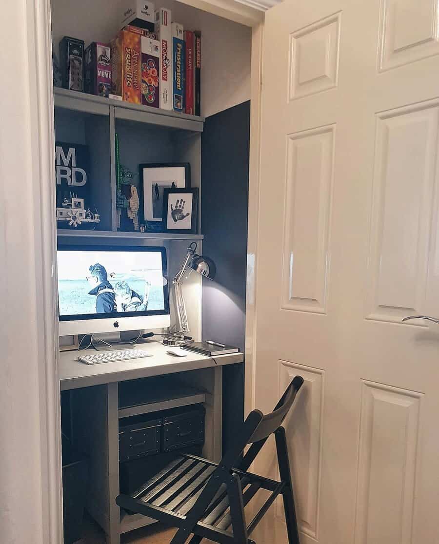 Small home office setup in a closet, featuring a computer desk, chair, shelves with board games, and a desktop computer with a lamp