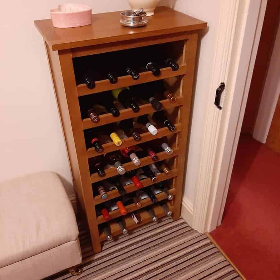 Wooden wine rack filled with bottles, placed against a wall, next to a cushioned seat on a striped carpet