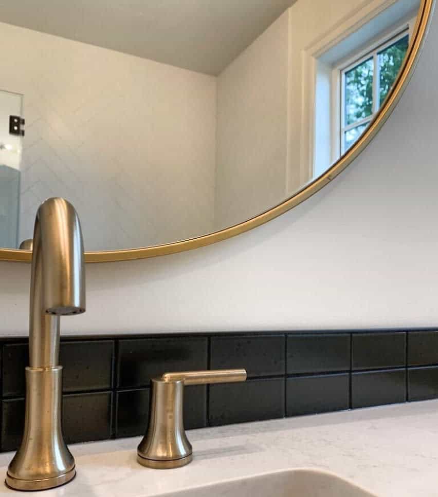 Elegant bathroom with black tile backsplash, brushed gold fixtures, white countertop, and round mirror with gold trim.