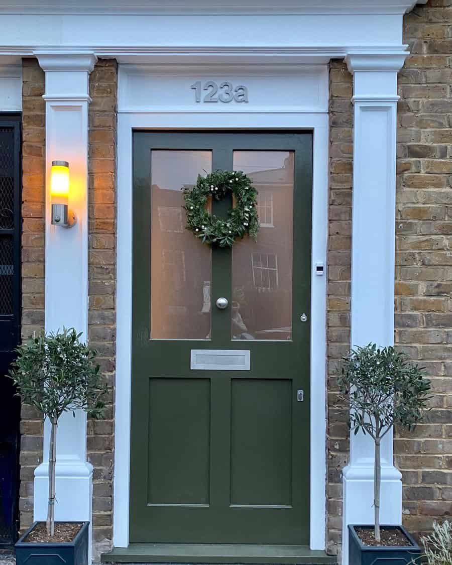 Green front door with a wreath, house number 123a, flanked by two small trees and a wall light, on a brick wall