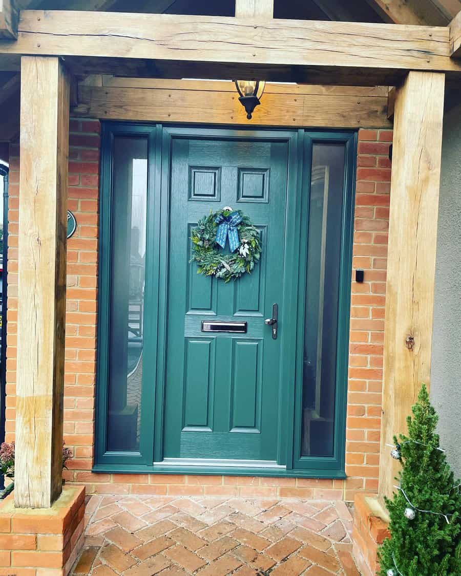 Green front door with a wreath under a wooden porch, brick wall and small fir tree in foreground