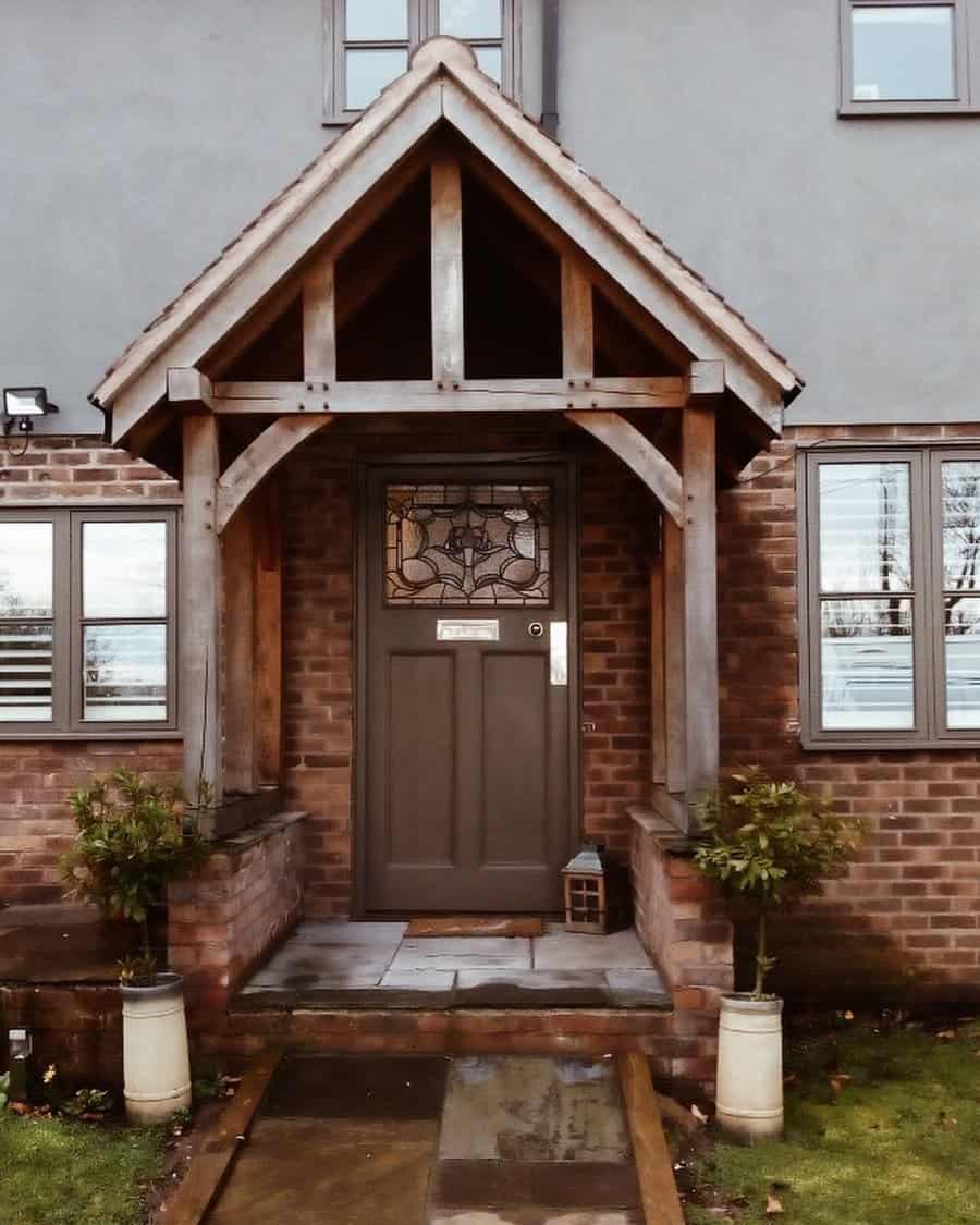 A wooden door with a decorative glass panel, surrounded by a brick facade and sheltered by a wooden porch, with two potted plants