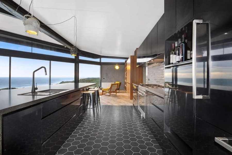 Modern kitchen with black cabinets, black hexagonal floor tiles, and large windows revealing an ocean view; dining area with wooden chairs