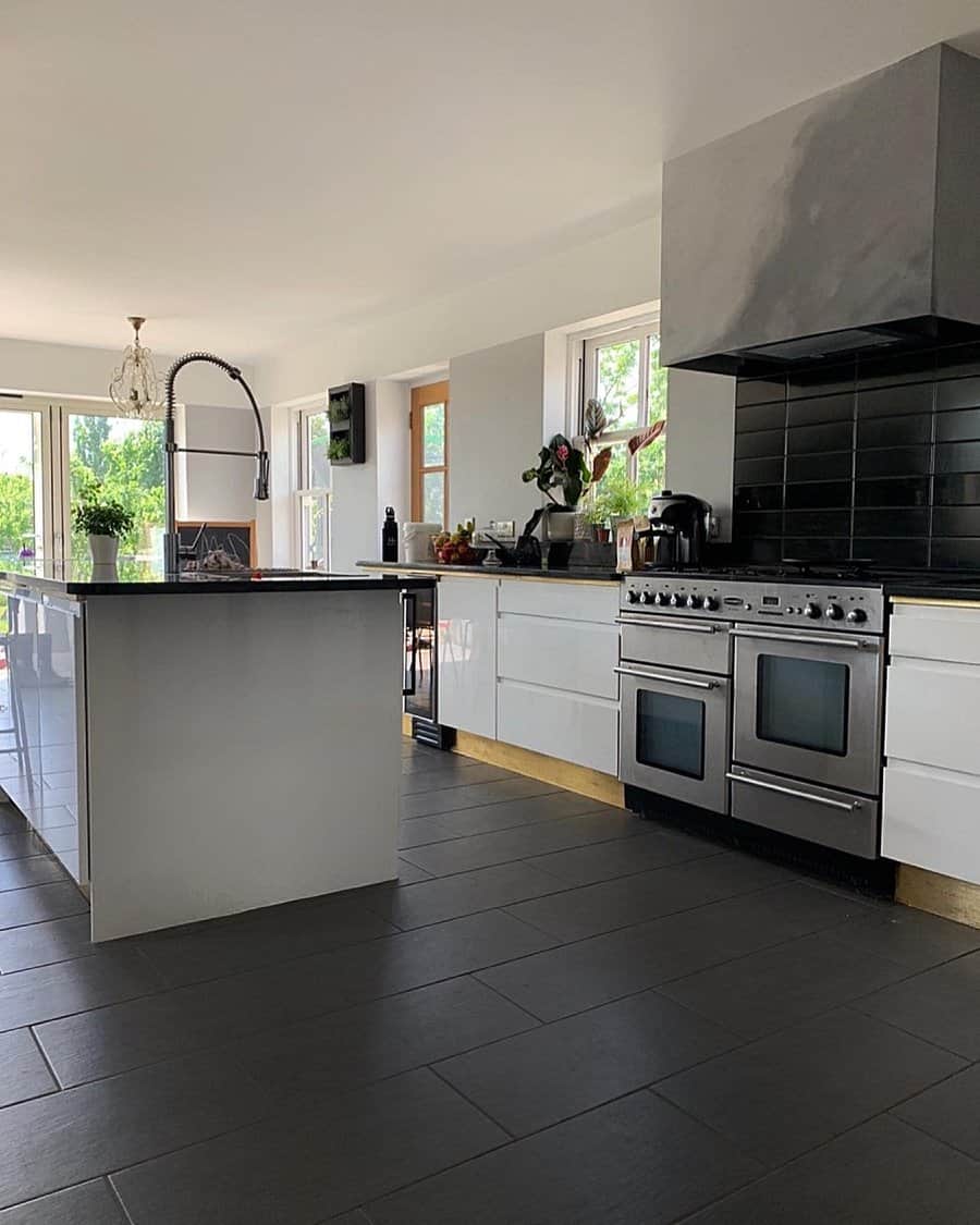 Modern kitchen with black tile floor, white cabinets, black backsplash, stainless steel stove, and island under chandelier; large windows