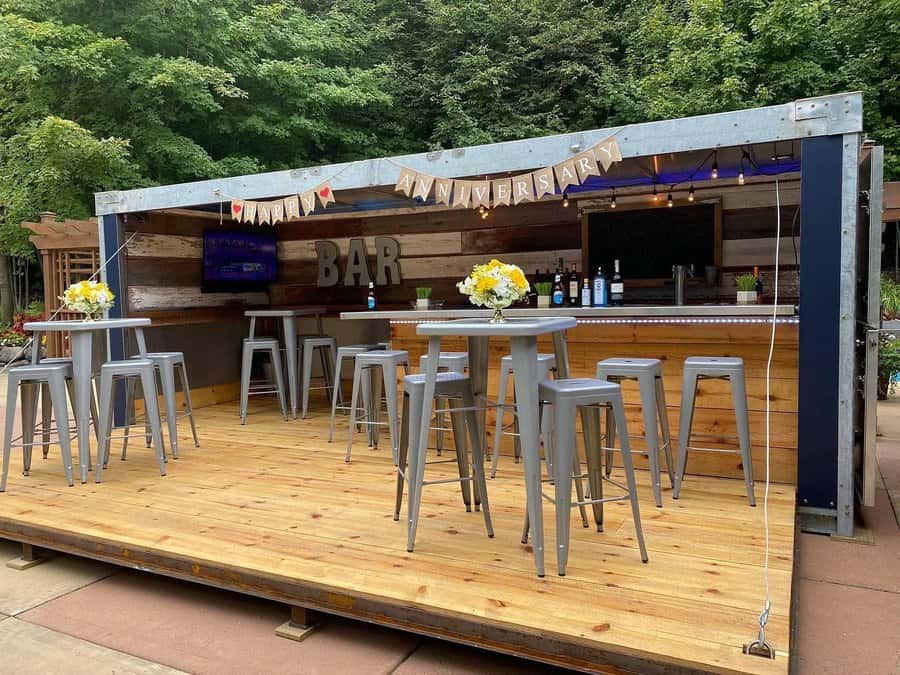 Outdoor bar setup with wooden deck, metal stools, floral decorations, and a sign reading "ANNIVERSARY" under a canopy