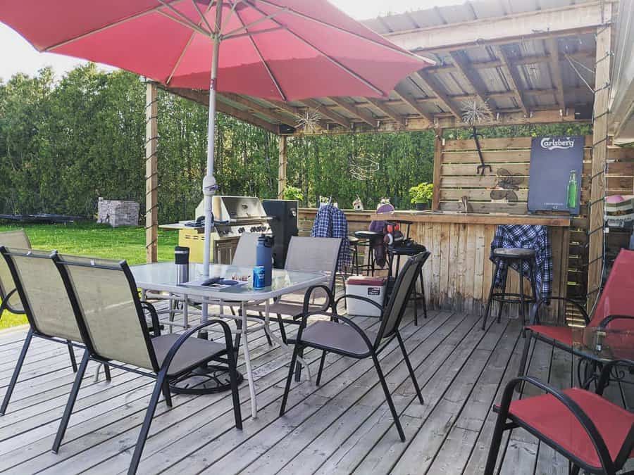 Outdoor deck with seating, table, umbrella, and a bar area under a pergola; grill and fridge visible; surrounded by greenery