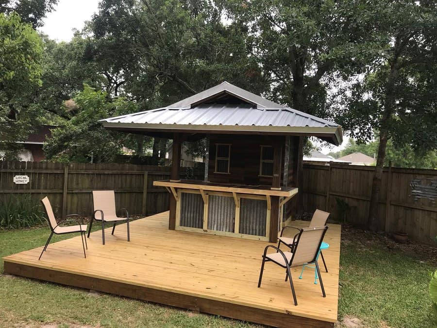 Wooden deck with a small hut, metal roof, and bar area, surrounded by trees and a fence, with several chairs on the deck