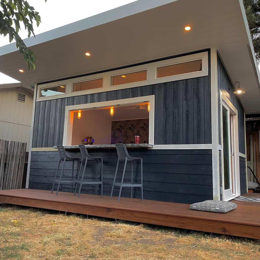 Modern outdoor bar area with two stools, a large serving window, and overhead lights; the deck is surrounded by greenery