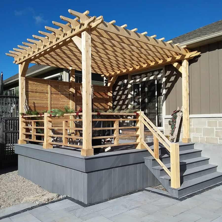 A wooden pergola with a lattice roof shades a small deck, featuring railings and steps, attached to a house 