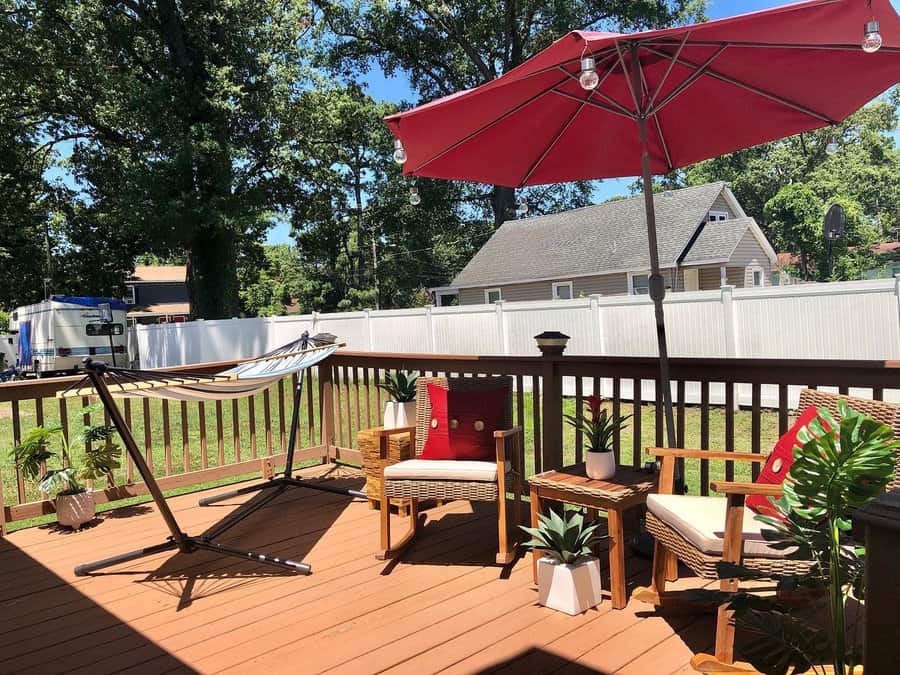 Sunny deck with two chairs, a red umbrella, a hammock, and potted plants. A white fence and trees are in the background