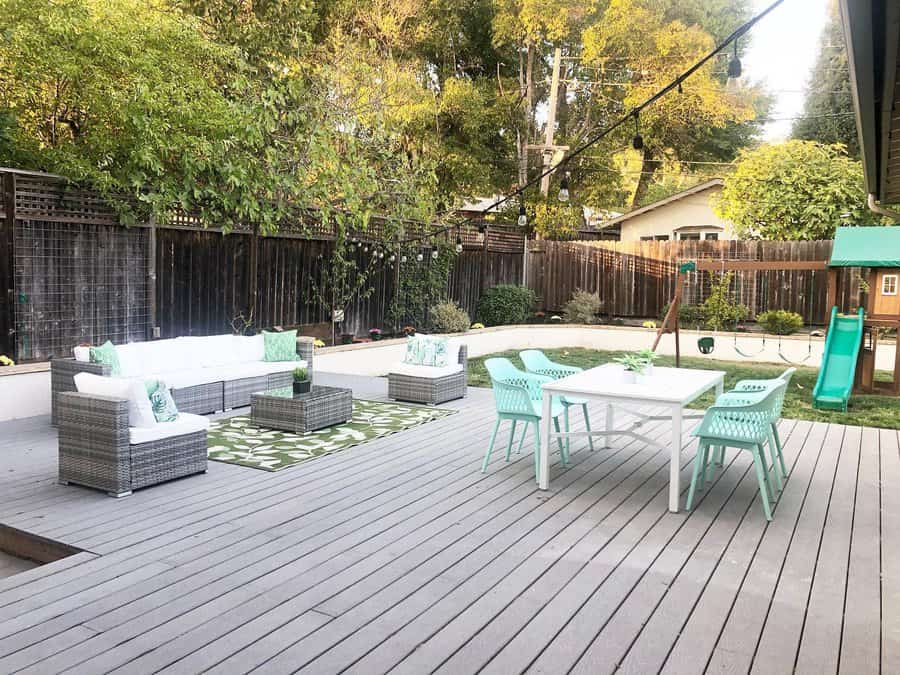 Spacious backyard with gray decking, white and gray patio furniture, turquoise chairs, a swing set, and green trees in the background