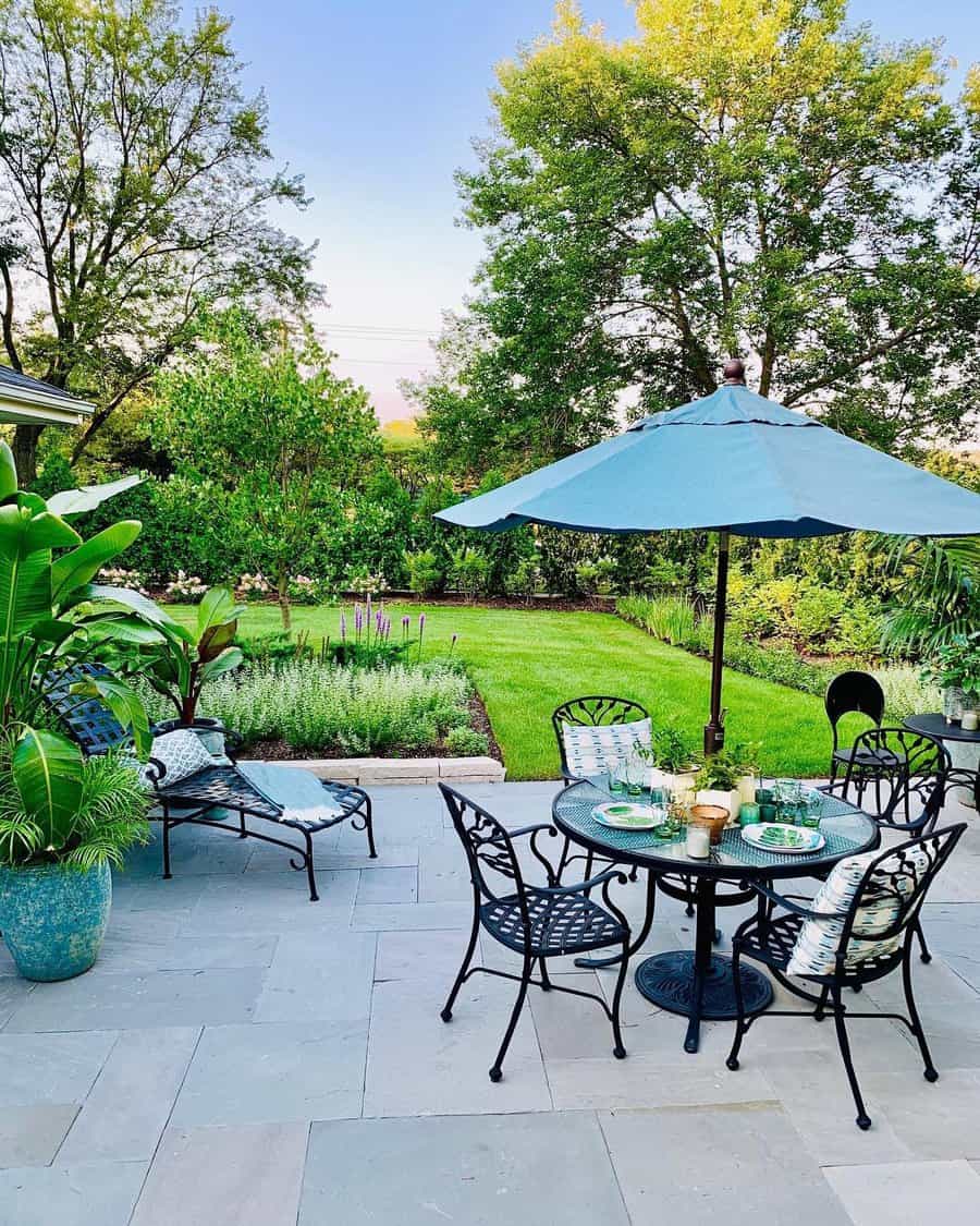 Backyard patio with black wrought-iron furniture, a round table set with dishes, an umbrella, and lush greenery in the background