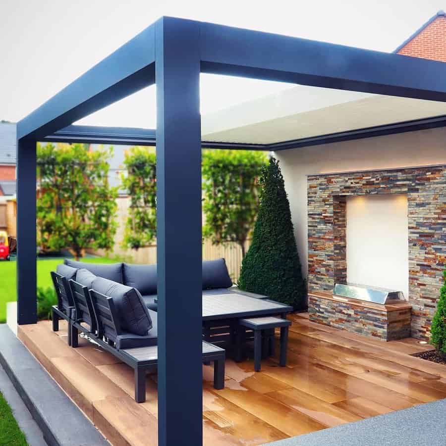 Modern outdoor patio with a metal pergola, wooden flooring, a dining set, and a stacked stone water feature, surrounded by greenery