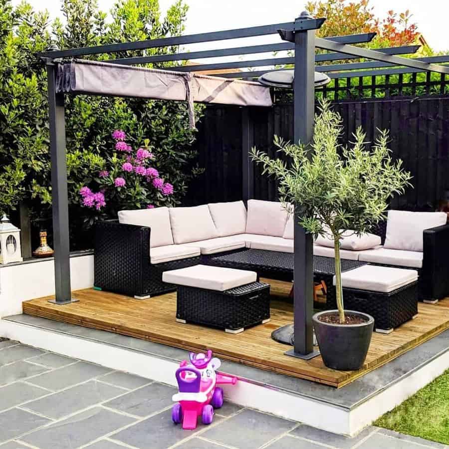 Patio with a black pergola, white cushioned seating, a potted tree, pink flowers, and a purple toy scooter on a tiled floor