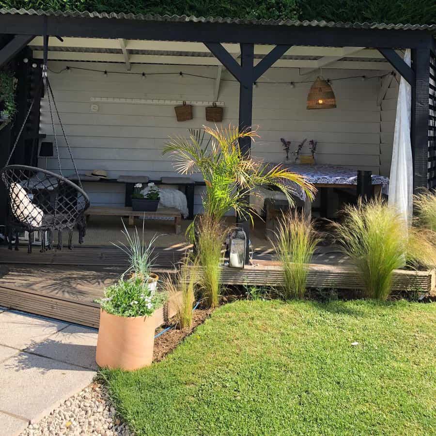 Outdoor patio with a covered lounge area, hanging chair, plants, and wooden decking, surrounded by a grassy lawn