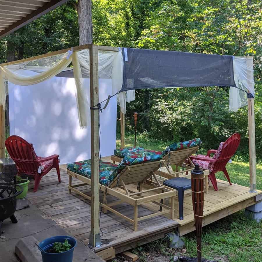 Outdoor seating area with wooden deck, chairs, and lounges featuring a screen and sheer fabric canopy for shade with greenery in the background