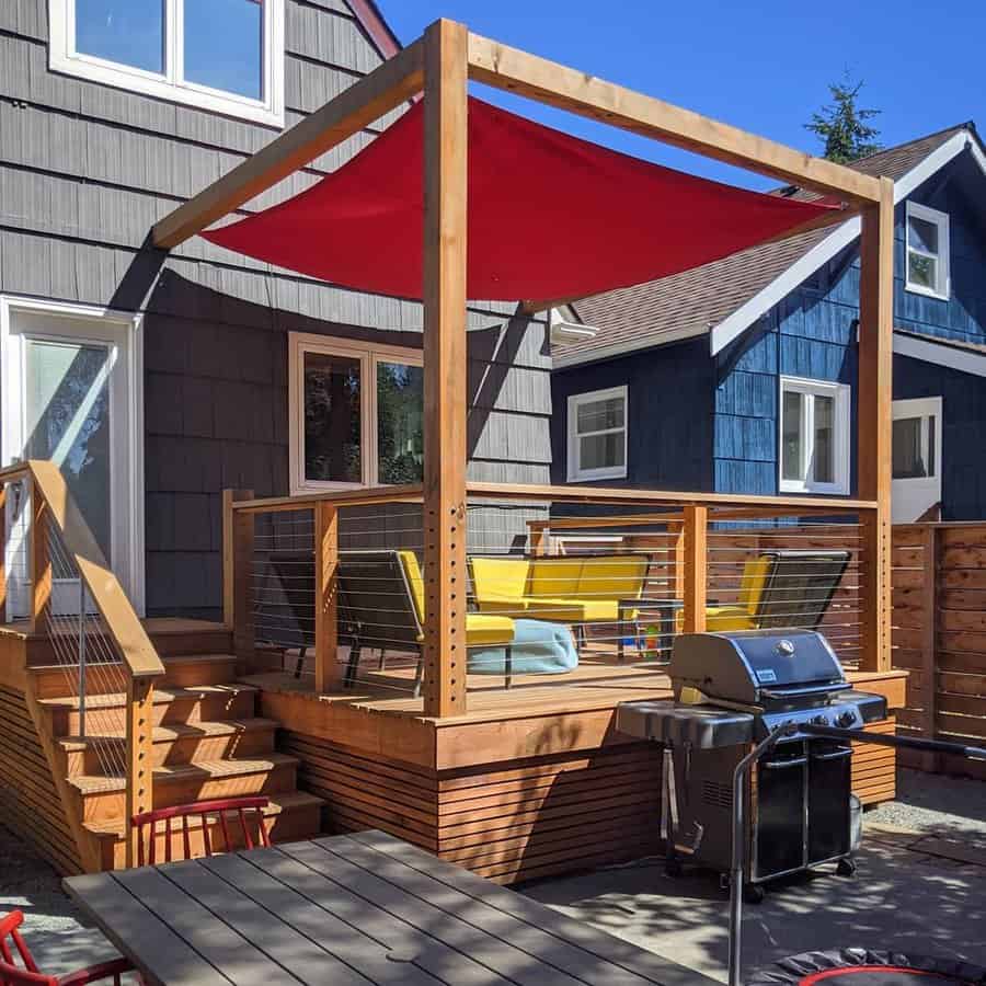 A wooden patio with a red shade sail, yellow seating, and a grill, adjacent house walls are gray and blue with white trim