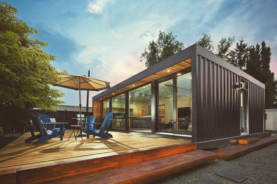 Modern container home with large windows, wooden deck, blue chairs, and umbrella on a sunny day, surrounded by trees