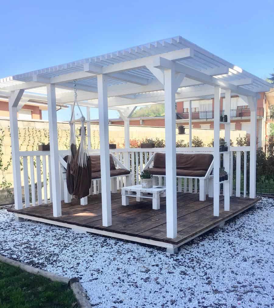 White pergola with swinging chairs and a small table on a wooden deck, surrounded by a gravel path