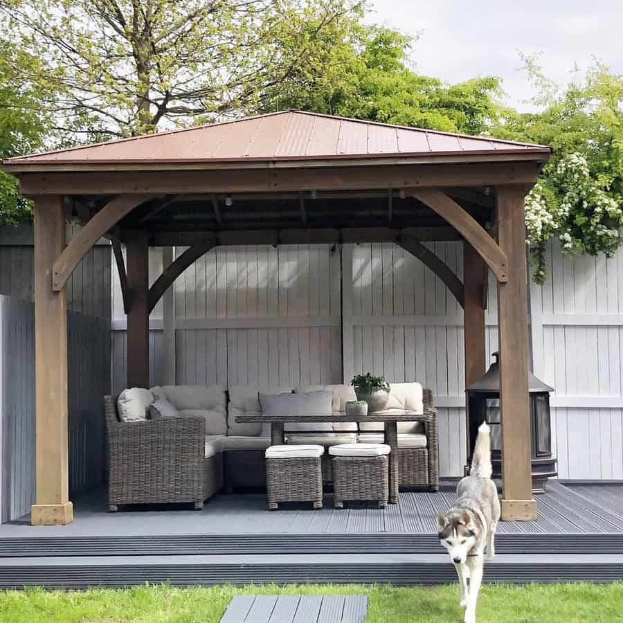 Cozy backyard gazebo with a cushioned wicker sectional, wooden dining table, and lantern on a raised deck, with a dog roaming nearby