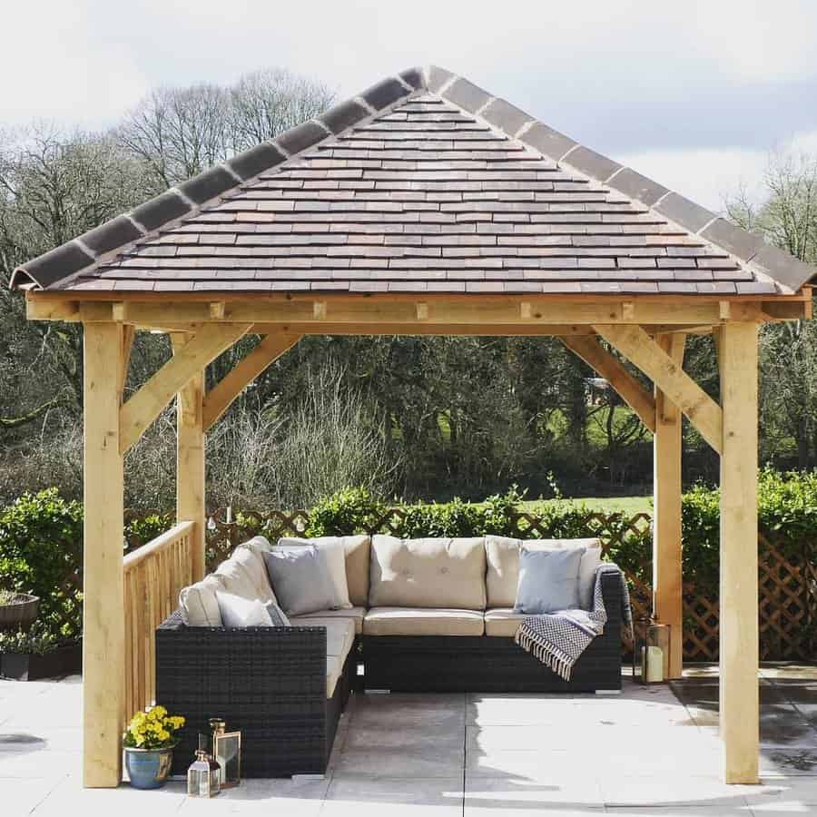 Wooden gazebo with a pitched roof, housing an L-shaped wicker sofa with cream cushions, surrounded by greenery and potted plants