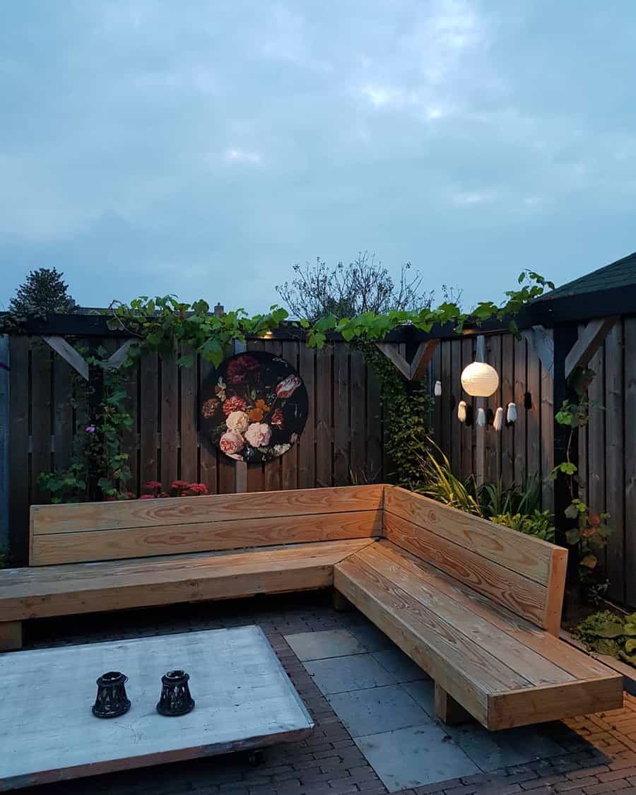 Cozy outdoor patio with wooden benches, lanterns, a floral wall art, and ambient lighting, set against a wooden fence with climbing plants