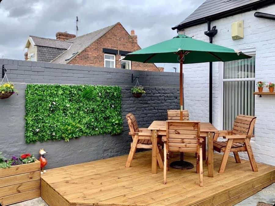 Cozy wooden deck with a patio table, chairs, and green umbrella; vertical garden on a gray brick wall, surrounded by potted plants