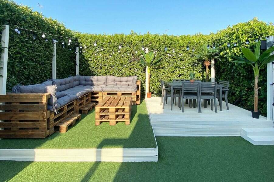 Modern outdoor lounge with pallet seating, gray cushions, a dining area, artificial grass, string lights, and lush green hedge walls