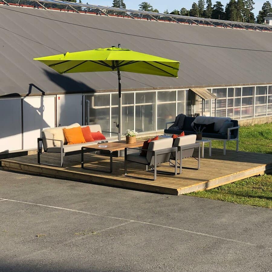 Outdoor wooden deck with sofas, colorful cushions, a coffee table, and a large green umbrella, set beside a building and lawn