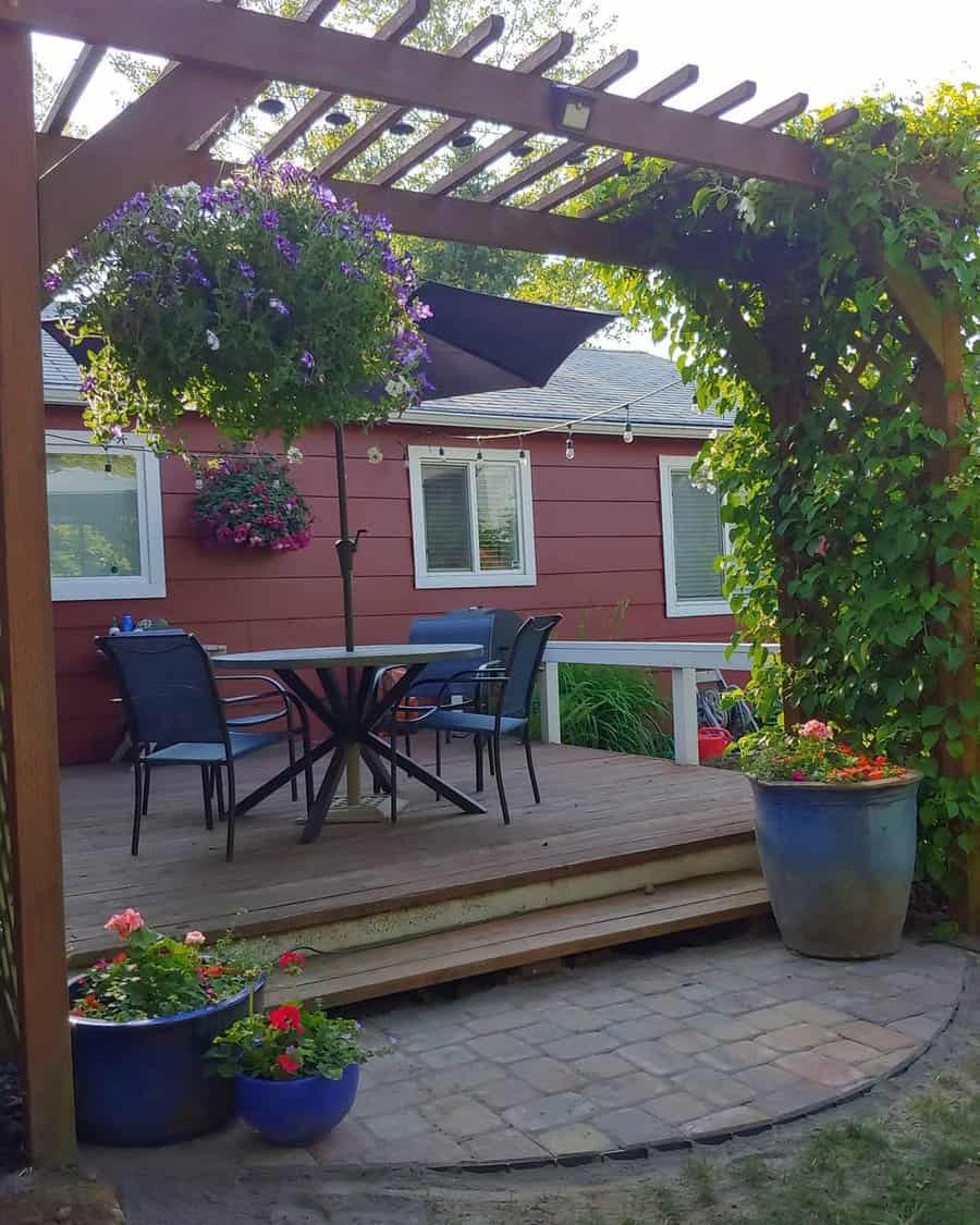 Wooden deck with patio table, chairs, and umbrella; surrounded by potted flowers and lush greenery under a pergola