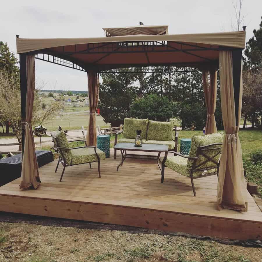 Gazebo with cushioned chairs and a table on a wooden deck, surrounded by greenery and an open grassy field in the background