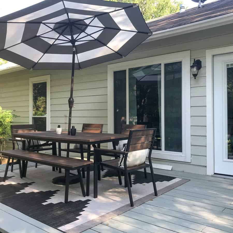 Outdoor patio with table, chairs, and bench under a striped umbrella on a rug; sliding glass doors lead to the house