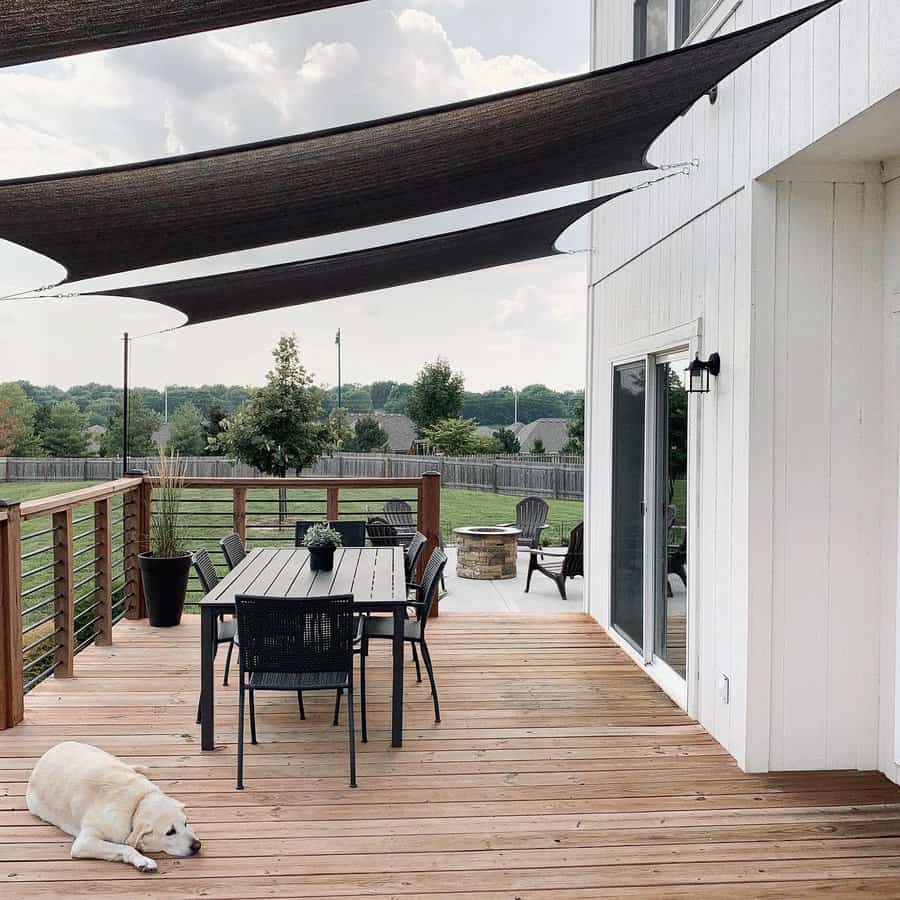 Modern backyard deck with black shade sails, a dining table, a fire pit, and a relaxed dog resting on the wooden floor