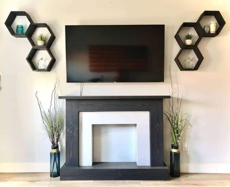 Wall-mounted TV above a dark fireplace, flanked by hexagonal shelves with plants and vases, two vases with tall grass on each side