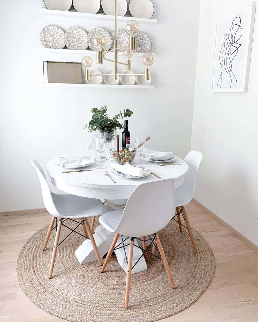 Minimalist dining area featuring a round white table, white chairs, a beige woven rug, and white shelves with plates; wall art is seen above