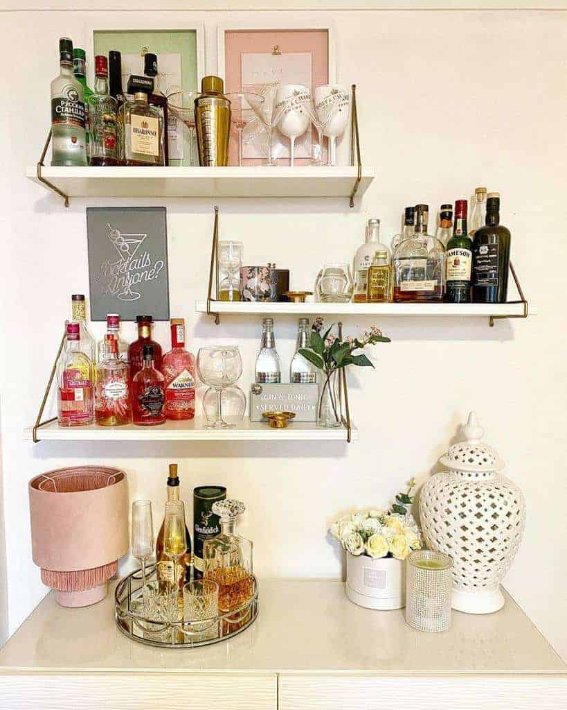 Stylish floating shelves with liquor bottles, glassware, and decor create a chic home bar setup with gold accents and soft pastel tones