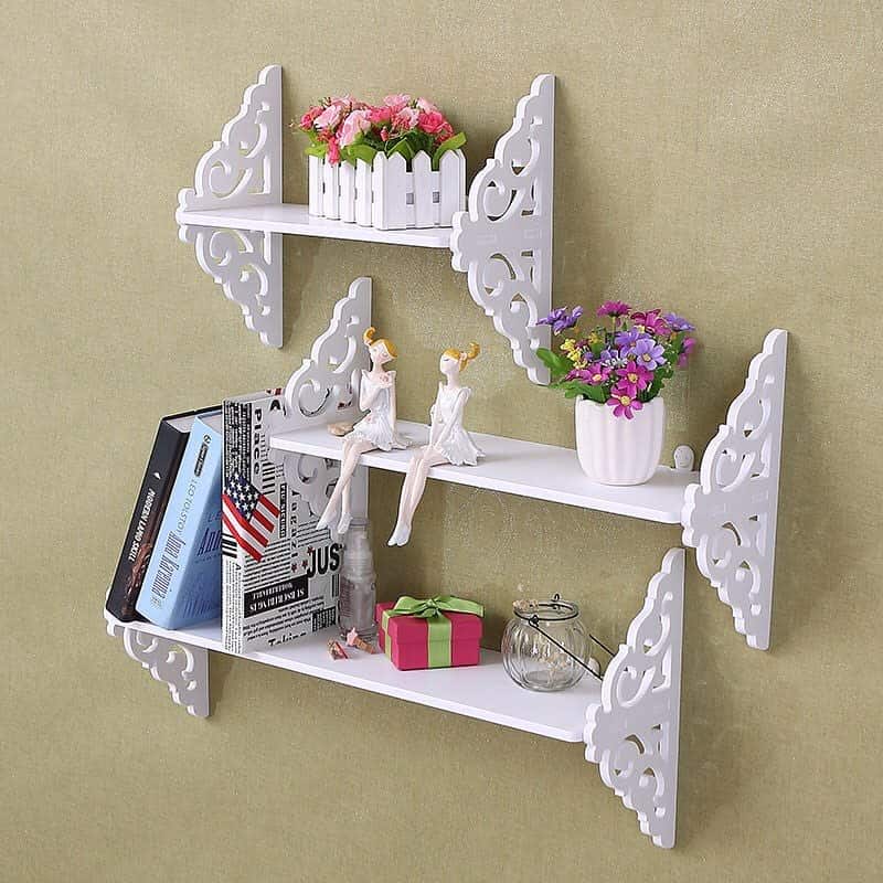 White decorative wall shelves with books, a gift box, flowers, and two figurines sitting on a middle shelf against a green wall