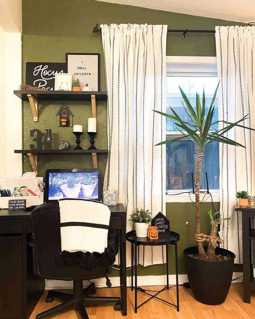 Home office with a desk, chair, and laptop; shelves hold decor and candles; a tall plant is beside the window with white curtains