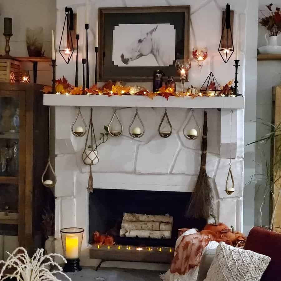 A cozy fireplace mantel decorated with fall leaves, hanging lanterns, candles, and a broom, creating a warm autumn atmosphere