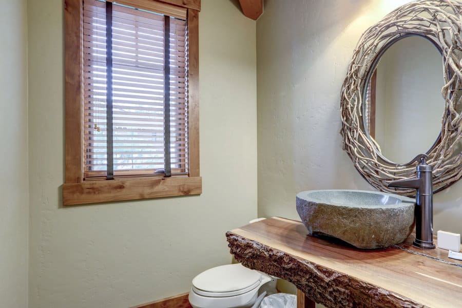 Bathroom with wooden accents, rustic sink on a live-edge countertop, round twig-framed mirror, and window with wooden blinds