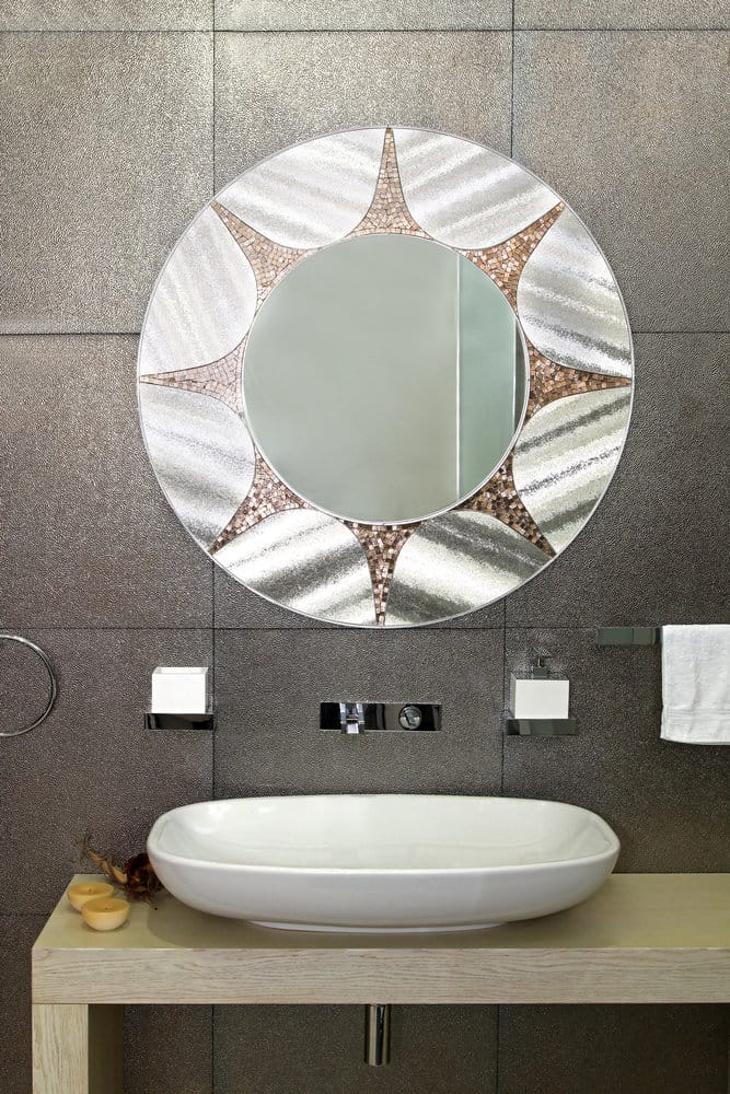 Bathroom with a metallic sun-themed mirror, white oval sink, and beige countertop against textured brown wall tiles