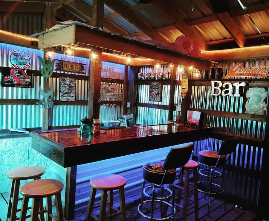 Cozy home bar with colorful LED lights, wooden stools, and eclectic decor, including signs and bottles on shelves