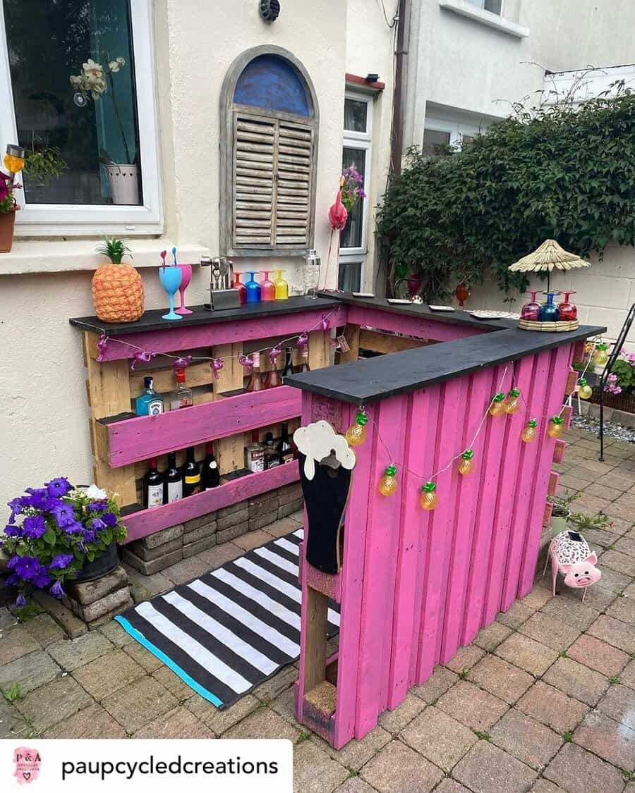 Colorful homemade outdoor bar with pink pallet walls, cocktails, and decorations; plants and drinks are placed on the counter