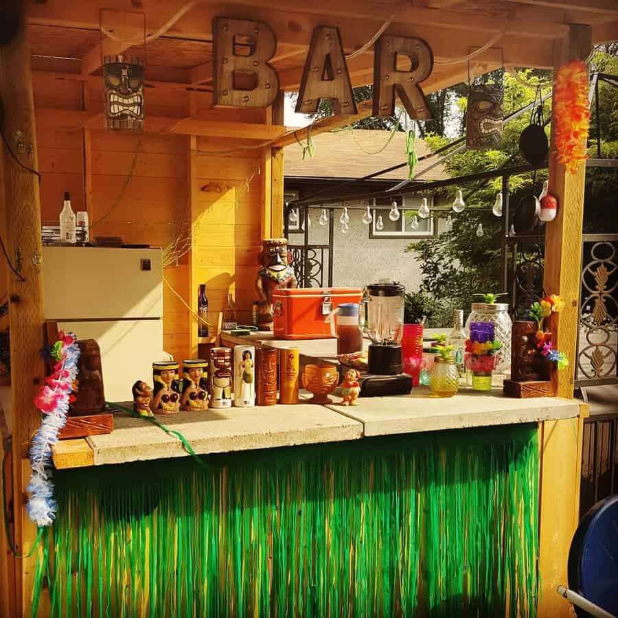 Outdoor tiki bar with a green fringe, tropical decor, colorful leis, various cups, and a blender with a "BAR" sign overhead