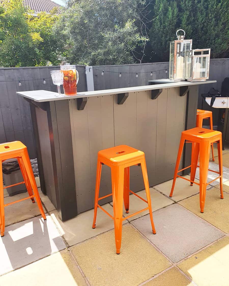 Outdoor bar setup with a black counter and three bright orange stools on a patio, drink pitcher and lanterns on the bar top